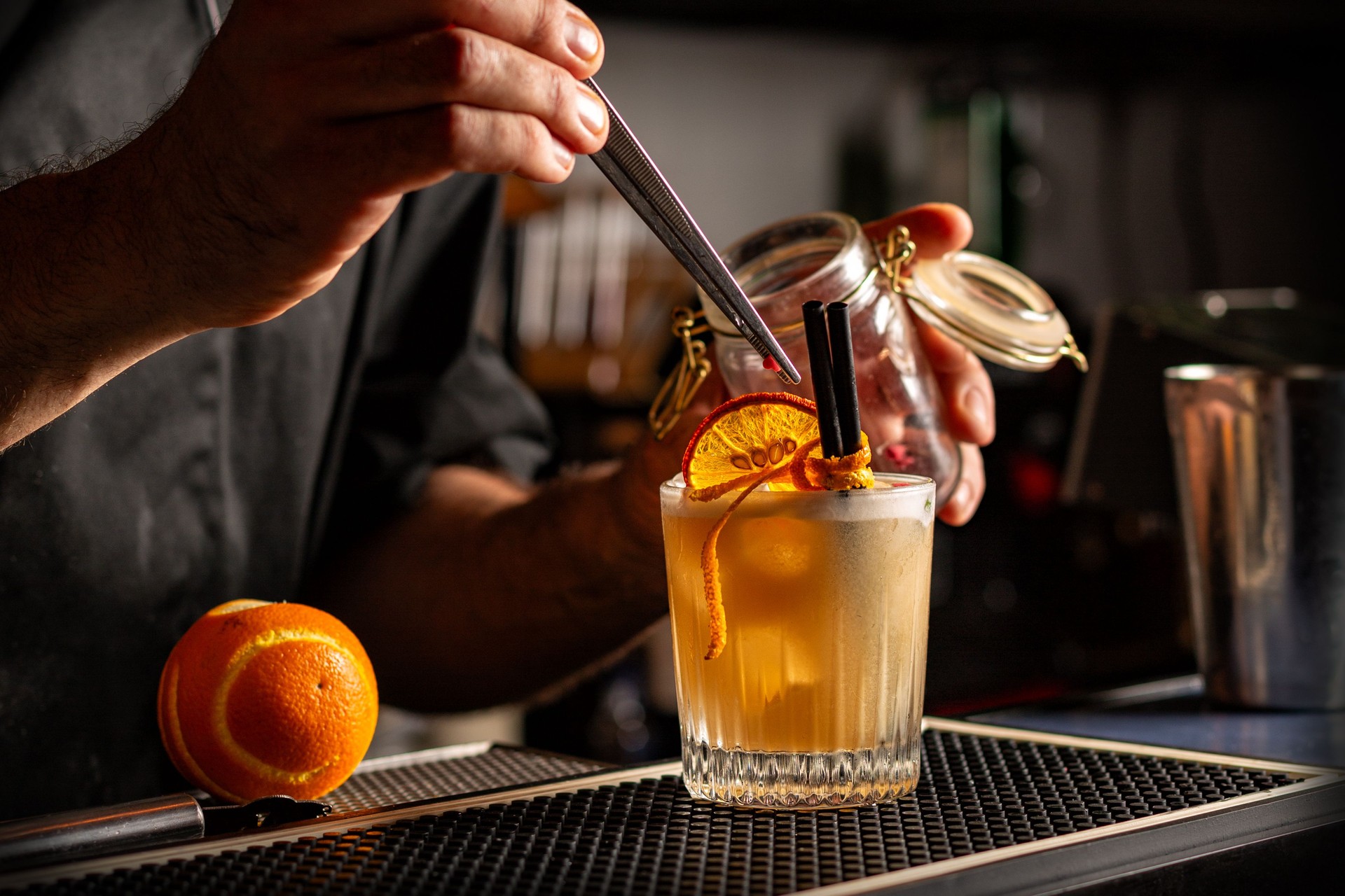 Preparing Smoky Spiced Orange Cocktail decorated with Dehydrated Orange Slices anf fresh orange peel. Bar or pub interior.